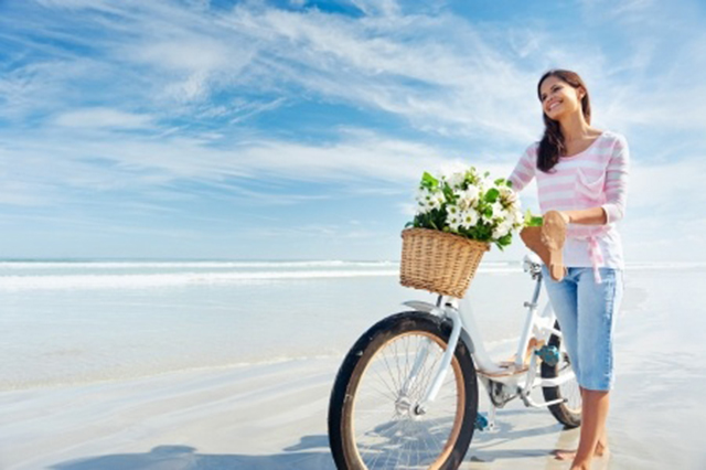 Image of young lady with bicycle walking along sands with high white clouds above beach horizon